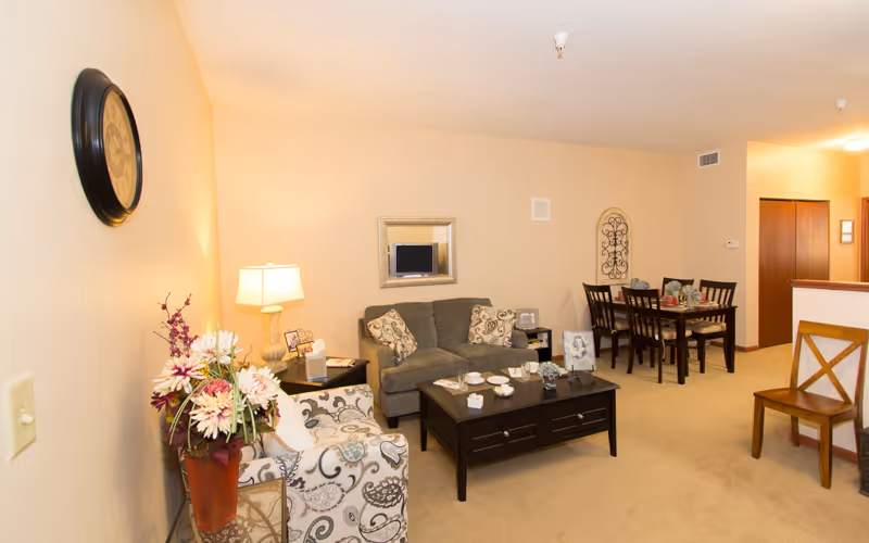 A cozy living room area with a gray sofa adorned with patterned pillows, a dark wooden coffee table with decorative items, and a patterned armchair next to a side table with a lamp and a vase of flowers. In the background, there is a dining area with a dark wooden dining table and chairs. The walls are light beige with a clock, a mirror, and decorative wall art.