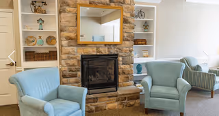 A cozy living room area featuring two light blue armchairs and a striped sofa arranged around a stone fireplace with a wooden-framed mirror above it. Built-in white shelves flank the fireplace, displaying decorative items such as vases, baskets, and small sculptures. The room has a neutral color palette with beige walls and carpeted flooring.