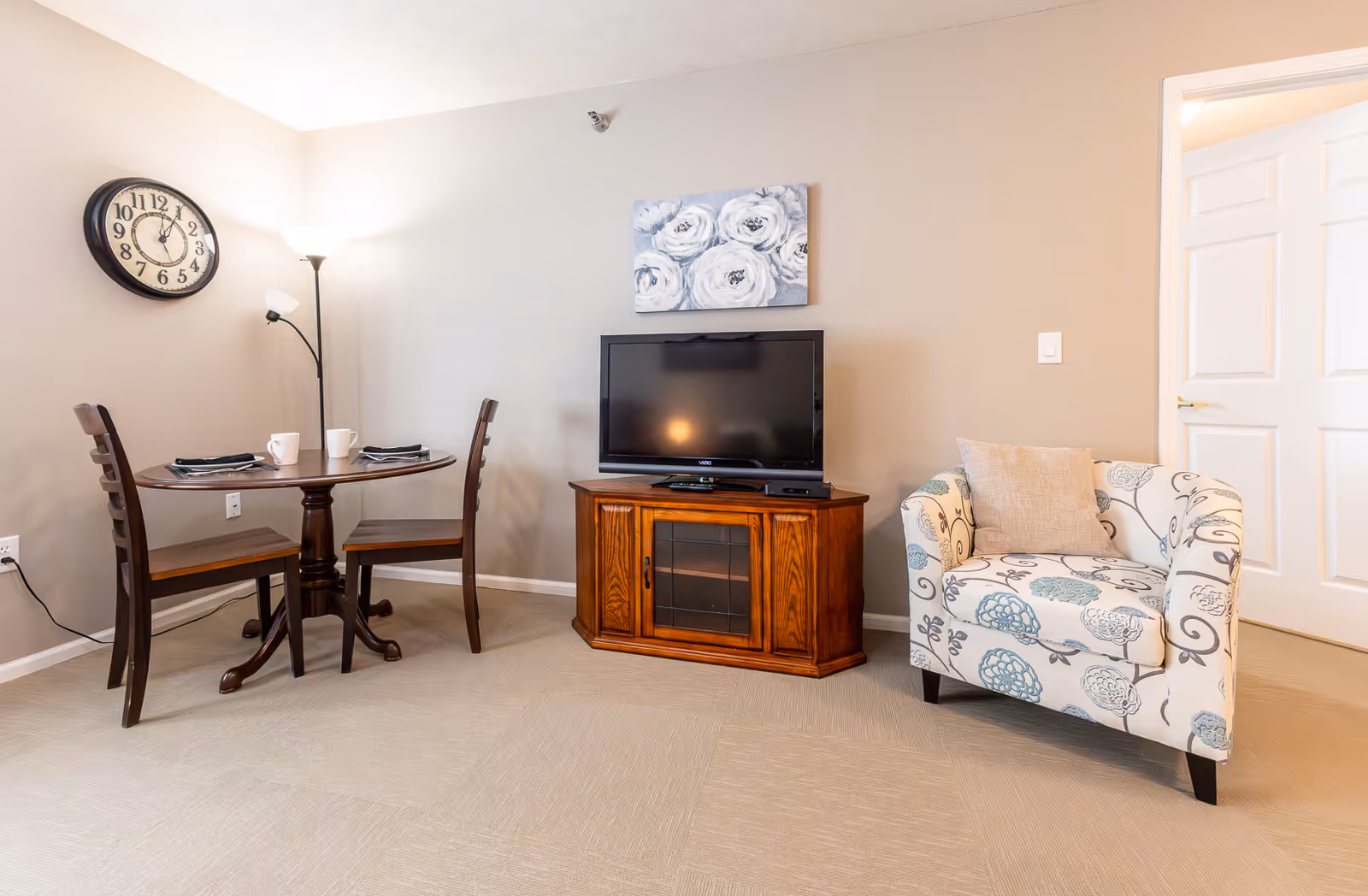 A cozy living area featuring a small round wooden dining table with two chairs, set with two cups and napkins. A floor lamp stands behind the table, illuminating the space. A wooden TV stand holds a flat-screen television, above which hangs a floral painting. To the right, there is a cushioned armchair with a floral pattern and a beige pillow. A white door is visible in the background.
