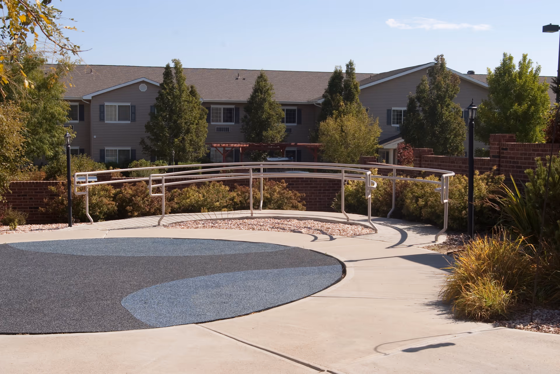 Outdoor area of a senior living facility with a curved concrete pathway featuring a blue and gray pattern, surrounded by bushes, trees, and lamp posts. In the background, there is a two-story building with multiple windows and a sloped roof.
