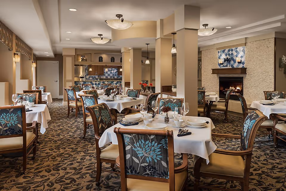 A cozy dining room with multiple round tables covered in white tablecloths, set with plates, bowls, glasses, and cutlery. The chairs have wooden frames with floral patterned upholstery. The room features a carpet with a dark floral pattern, beige walls, and a fireplace with a blue and white tiled design above it. Pendant lights hang from the ceiling, and there is a kitchen area visible in the background with blue and white tiled backsplash.