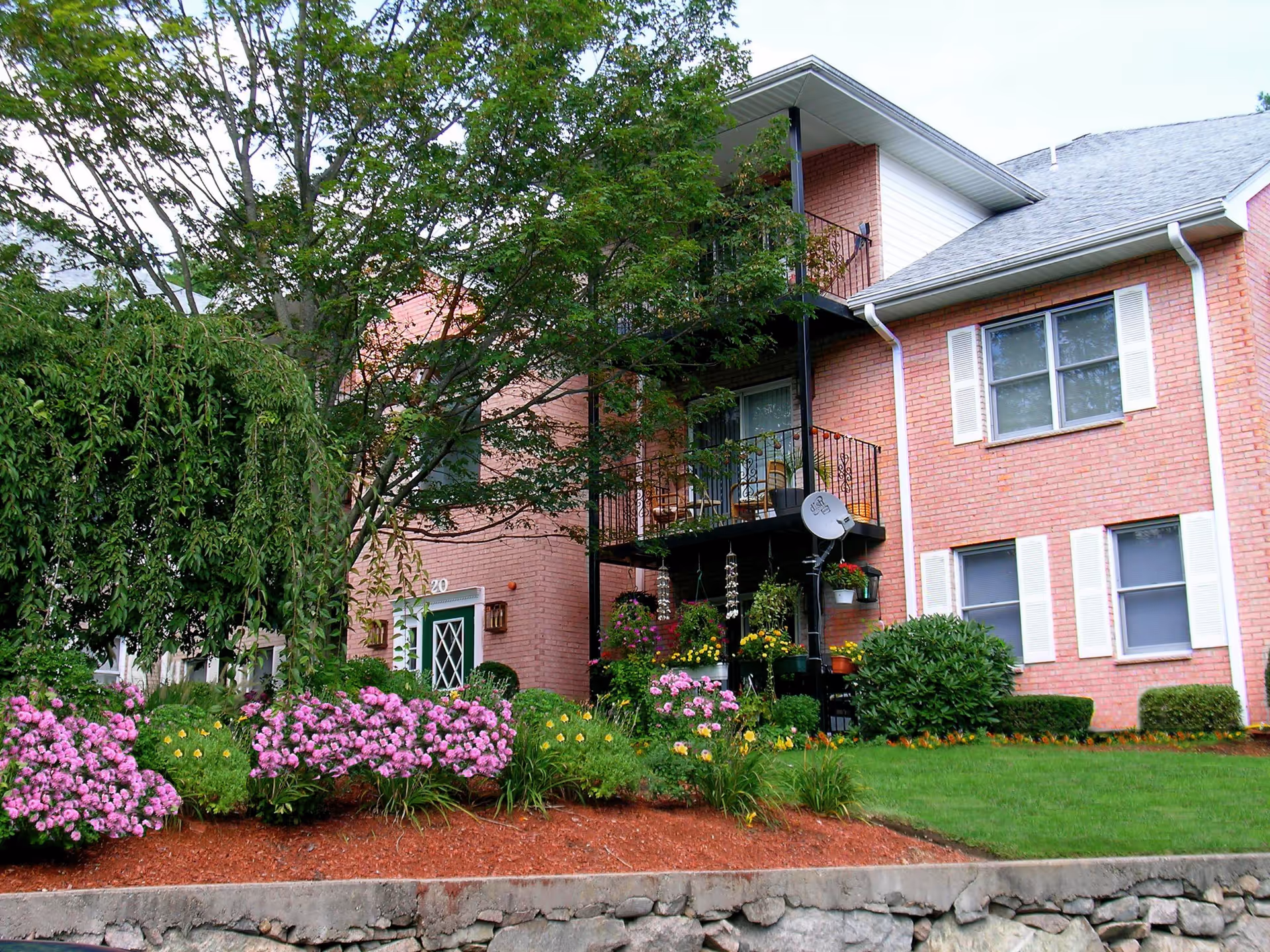 Exterior view of a two-story brick building with white window shutters, a balcony with outdoor chairs, and a satellite dish. The building is surrounded by green trees, bushes, and colorful flower beds with pink and yellow flowers. There is a stone retaining wall in the foreground.