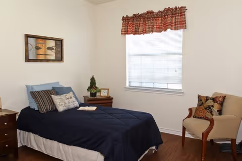 A simple furnished bedroom with a single bed, nightstands, a window with blinds and a plaid valance, and an upholstered chair.