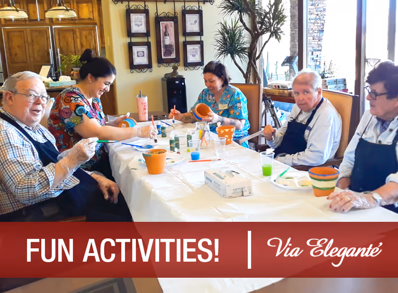 A group of elderly individuals and caregivers sitting around a table engaging in a pottery painting activity. They are smiling and wearing aprons and gloves. The table is covered with a white cloth and has paint supplies and small flower pots. The setting appears to be a cozy indoor room with wooden cabinets and decorative wall art in the background.
