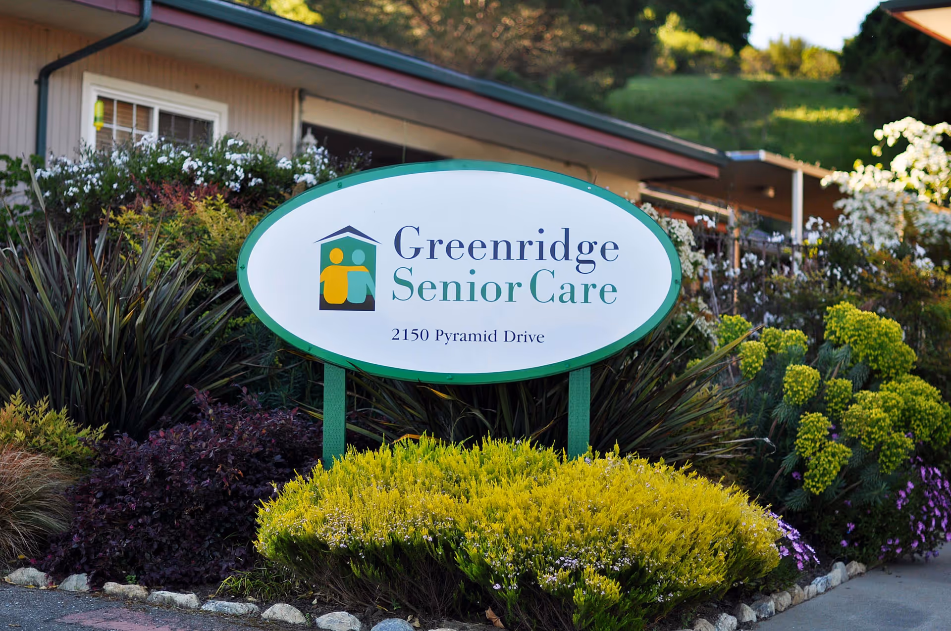 Outdoor view of a sign for Greenridge Senior Care located at 2150 Pyramid Drive, surrounded by various green and flowering plants with a building partially visible in the background.