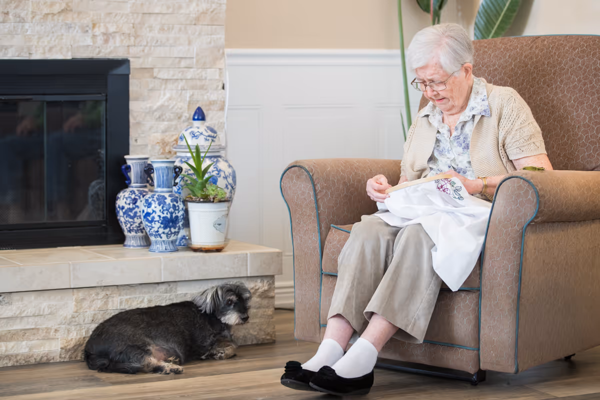 An elderly woman sitting in a comfortable armchair, engaged in embroidery or needlework. A small black and gray dog lies on the floor nearby. The setting includes a stone fireplace with decorative blue and white vases and a potted plant on the hearth.