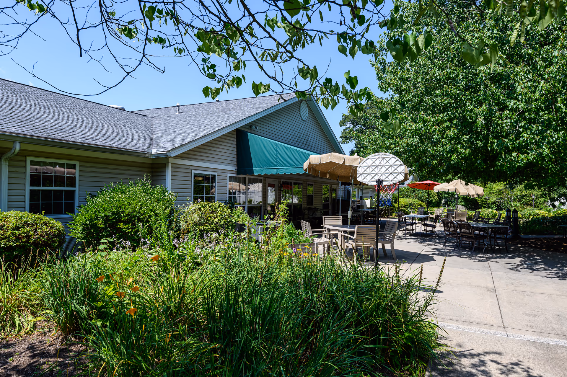 Outdoor patio area at Arden Courts - ProMedica Memory Care Community (Farmington) featuring multiple tables and chairs with umbrellas, surrounded by greenery and trees under a clear blue sky.
