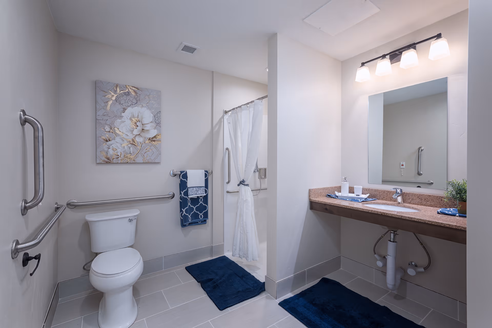 A clean and accessible bathroom with a white toilet, grab bars on the walls, a walk-in shower with a white curtain, a large mirror above a countertop sink, blue bath mats on the floor, and a decorative floral painting on the wall.