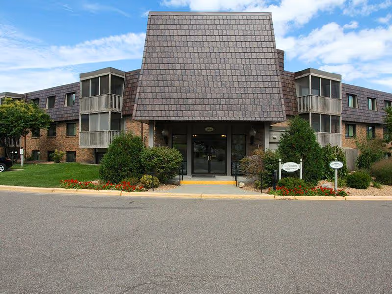 Front entrance of a low-rise brick senior living building with a large shingled entry canopy, landscaped beds, and screened balconies.