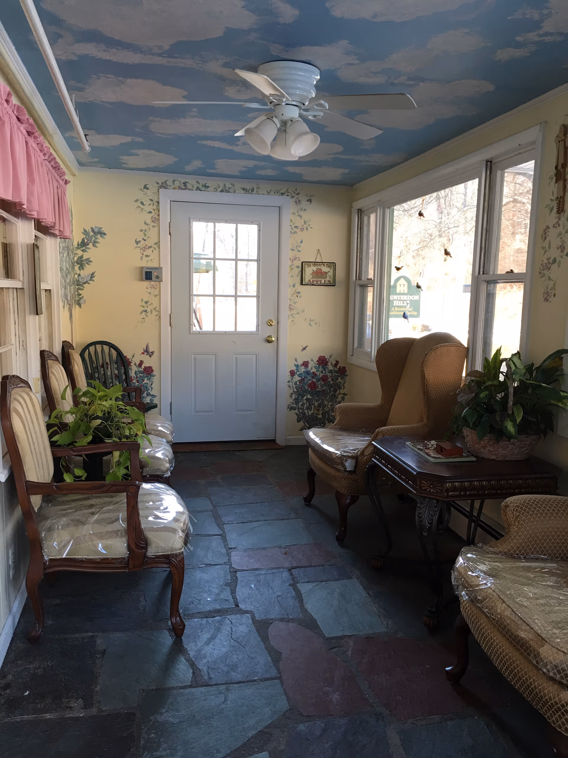 A cozy indoor sitting area with a stone tile floor, several chairs with plastic covers, a small wooden table with a plant and books, and a door with windows letting in natural light. The ceiling is painted with a blue sky and white clouds, and the walls have floral decorations and a small sign that reads 'Apples'.