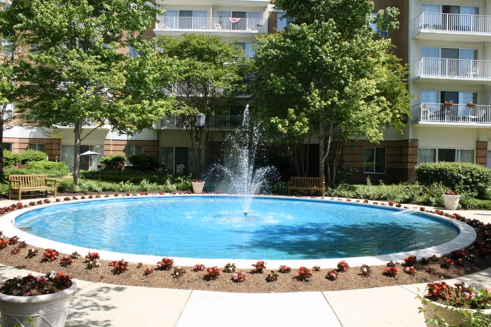 A circular water fountain with a central jet spraying water, surrounded by a flower bed with red flowers. There are benches and green trees around the fountain, with a multi-story building in the background featuring balconies and windows.