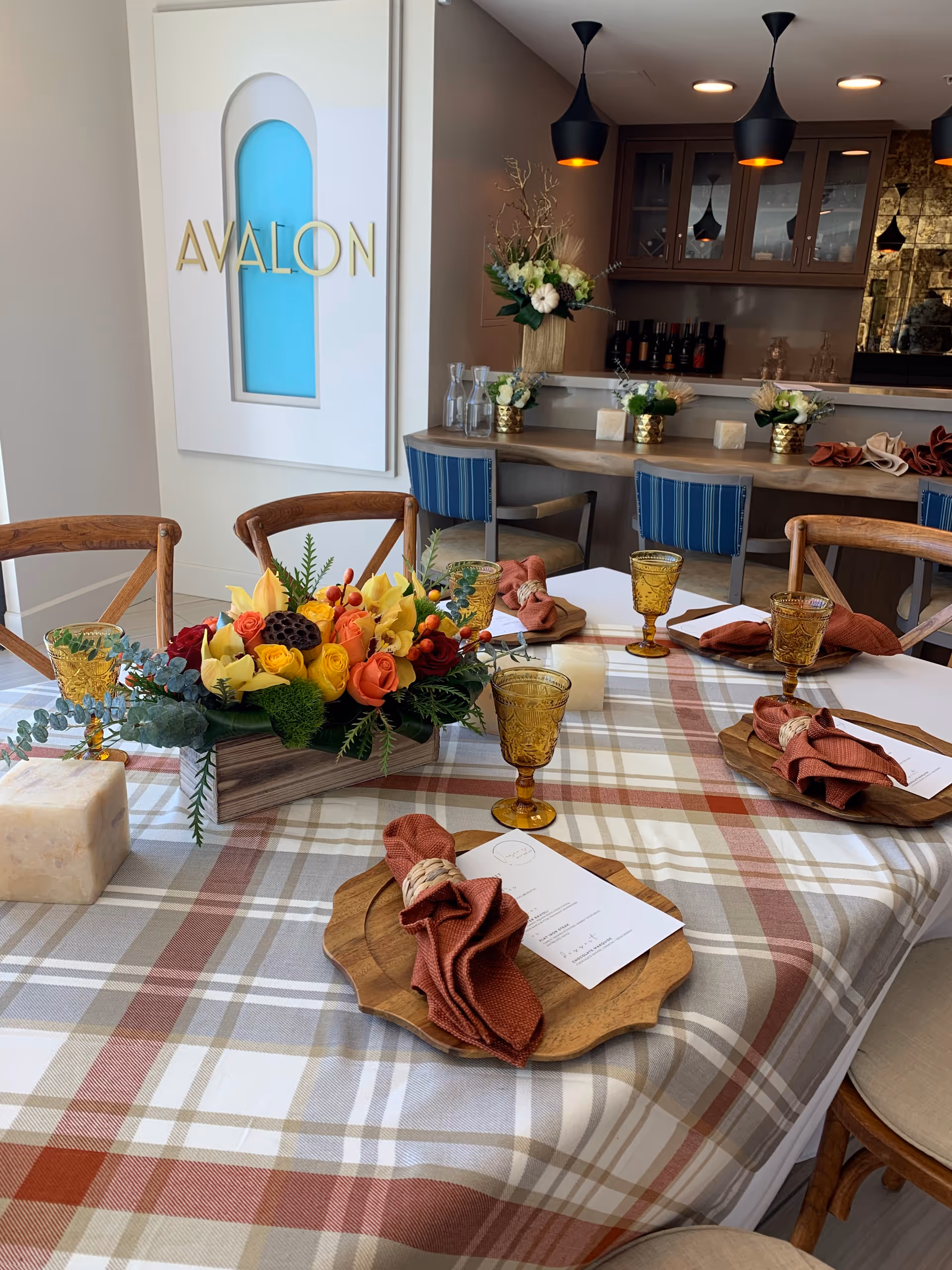 A dining table set with a plaid tablecloth, wooden plates, amber-colored goblets, and rust-colored cloth napkins. A centerpiece of vibrant flowers including yellow, red, and orange blooms is placed in the middle of the table. In the background, there is a bar area with blue chairs, decorative plants, and pendant lights hanging from the ceiling. A wall decoration with the word 'AVALON' is visible on the left side.