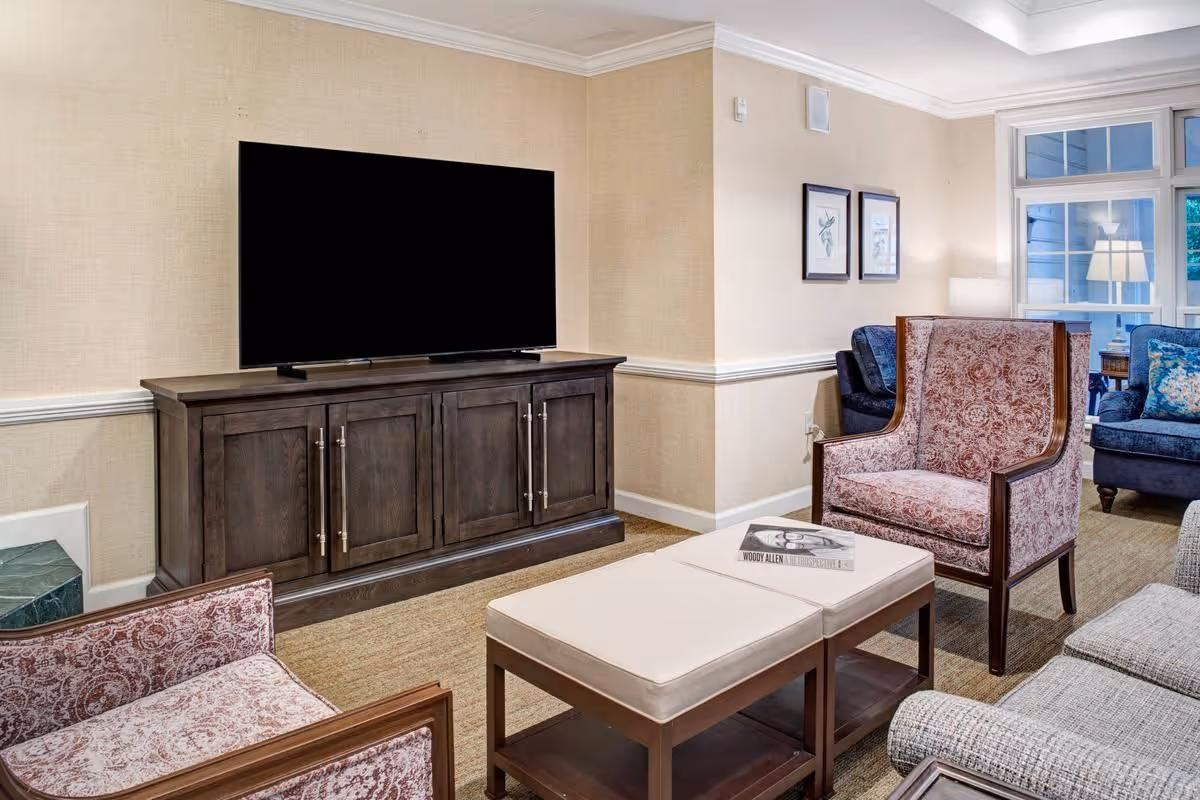 Communal living room with a large flat-screen TV on a dark wood cabinet, upholstered armchairs, an ottoman, and windows with a lamp.