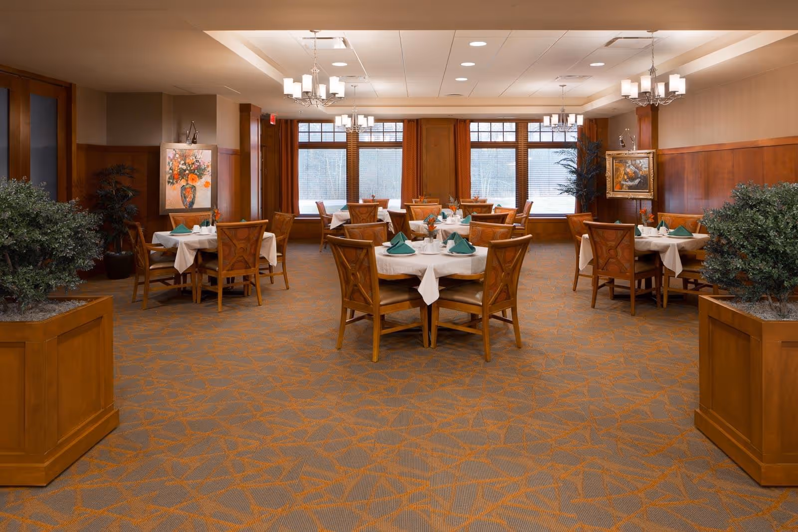 Spacious dining room with round tables set with white tablecloths and green napkins, wooden chairs and warm wood-paneled walls.