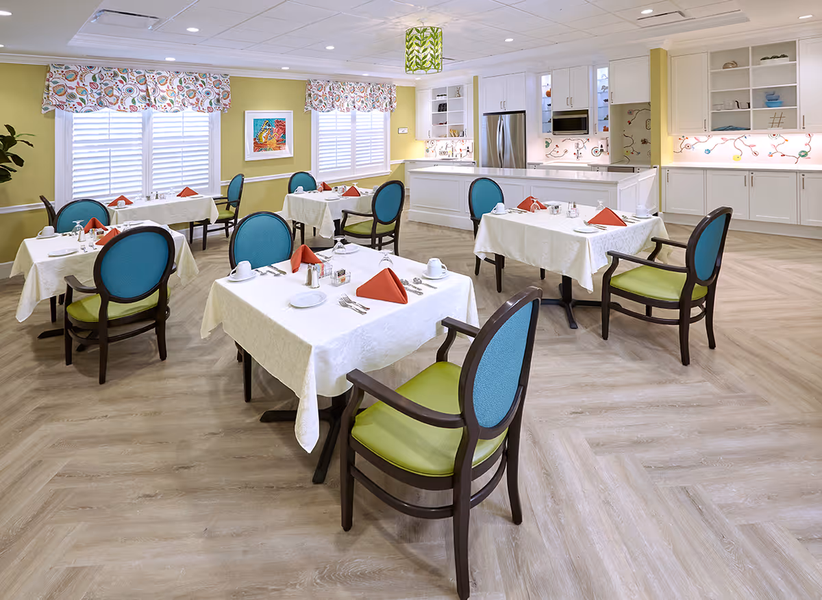 A bright and clean dining room with several tables covered in white tablecloths, each set with white plates, cups, silverware, and red folded napkins. The chairs have blue backs and green seats. The room has large windows with colorful patterned valances, light yellow walls, and a modern kitchen area with white cabinetry and stainless steel appliances in the background.