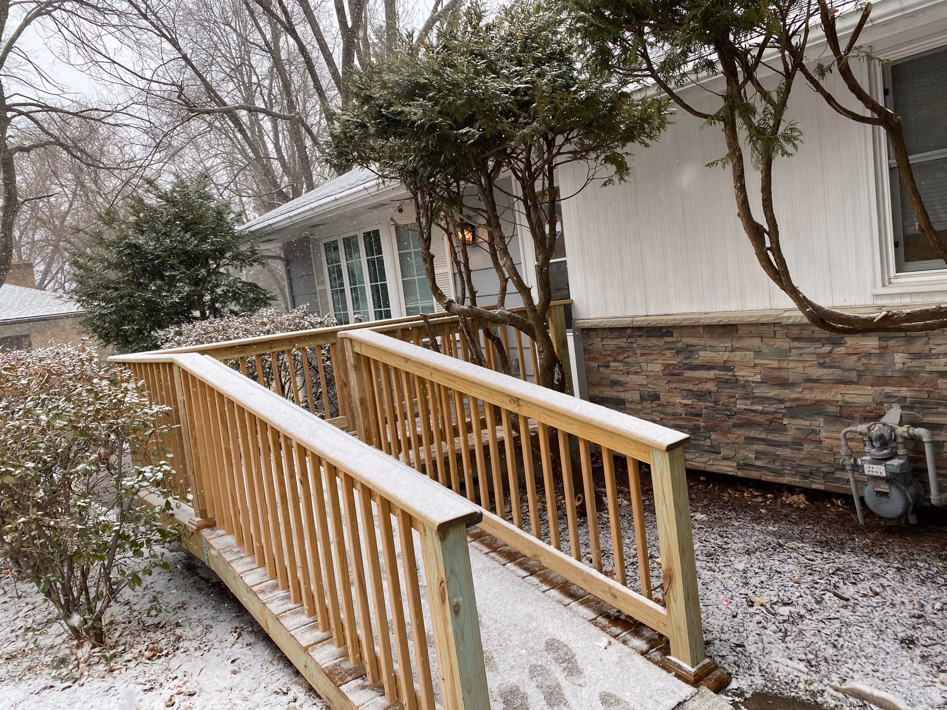 Snow-dusted wooden accessibility ramp leading to the front entrance of a single-story house with shrubs and trees.