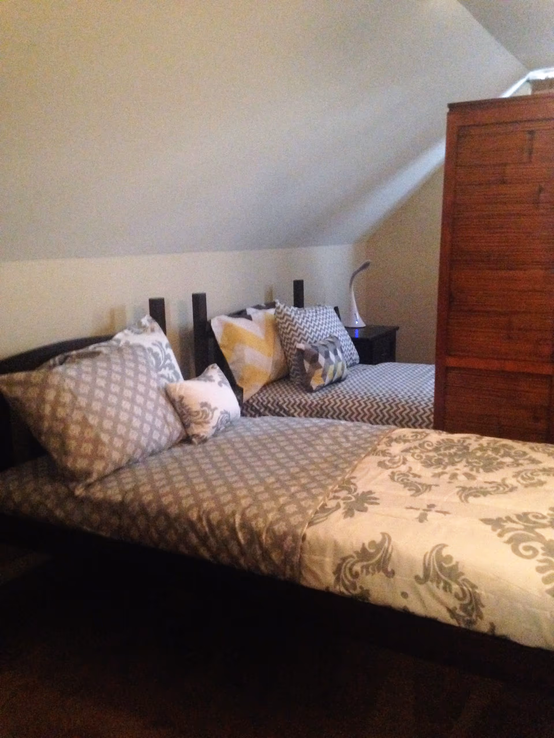 A bedroom with two single beds placed side by side, each made with patterned bedding and multiple pillows. There is a wooden wardrobe on the right side and a small nightstand with a modern lamp between the beds. The room has slanted ceilings and neutral-colored walls.