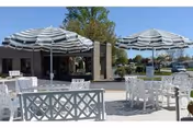Outdoor patio with white tables and chairs shaded by striped umbrellas in front of a low building.