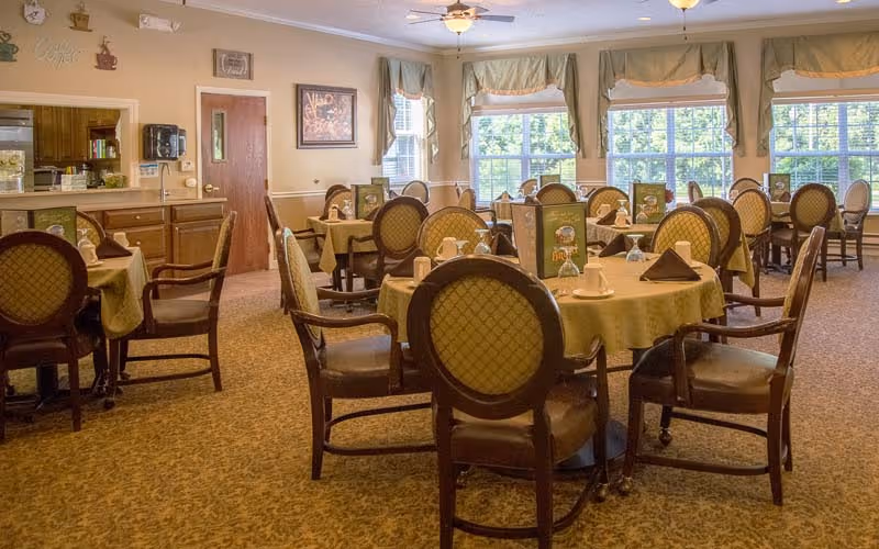 A dining room with multiple round tables covered with yellow tablecloths, each set with cups, napkins, and menus. The room has large windows with green curtains allowing natural light to fill the space. There are wooden chairs with cushioned seats and backs around each table. The walls are decorated with framed pictures and coffee-themed wall art. Ceiling fans and light fixtures are visible on the ceiling.