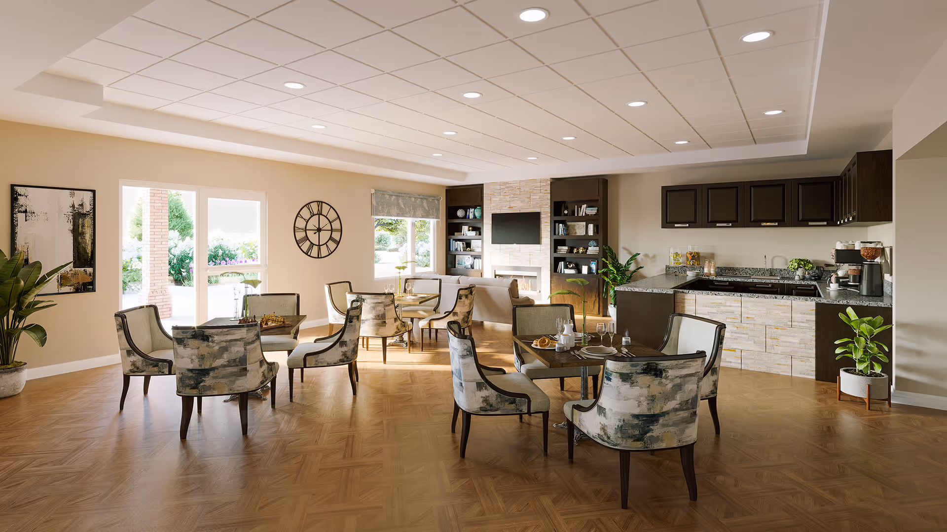 A spacious and well-lit senior living common area featuring multiple seating arrangements with upholstered chairs around small tables. The room has large windows and glass doors letting in natural light, a wall clock, indoor plants, and a kitchenette with dark cabinets and a granite countertop. There is also a cozy sitting area with a fireplace and built-in shelves with decorative items.