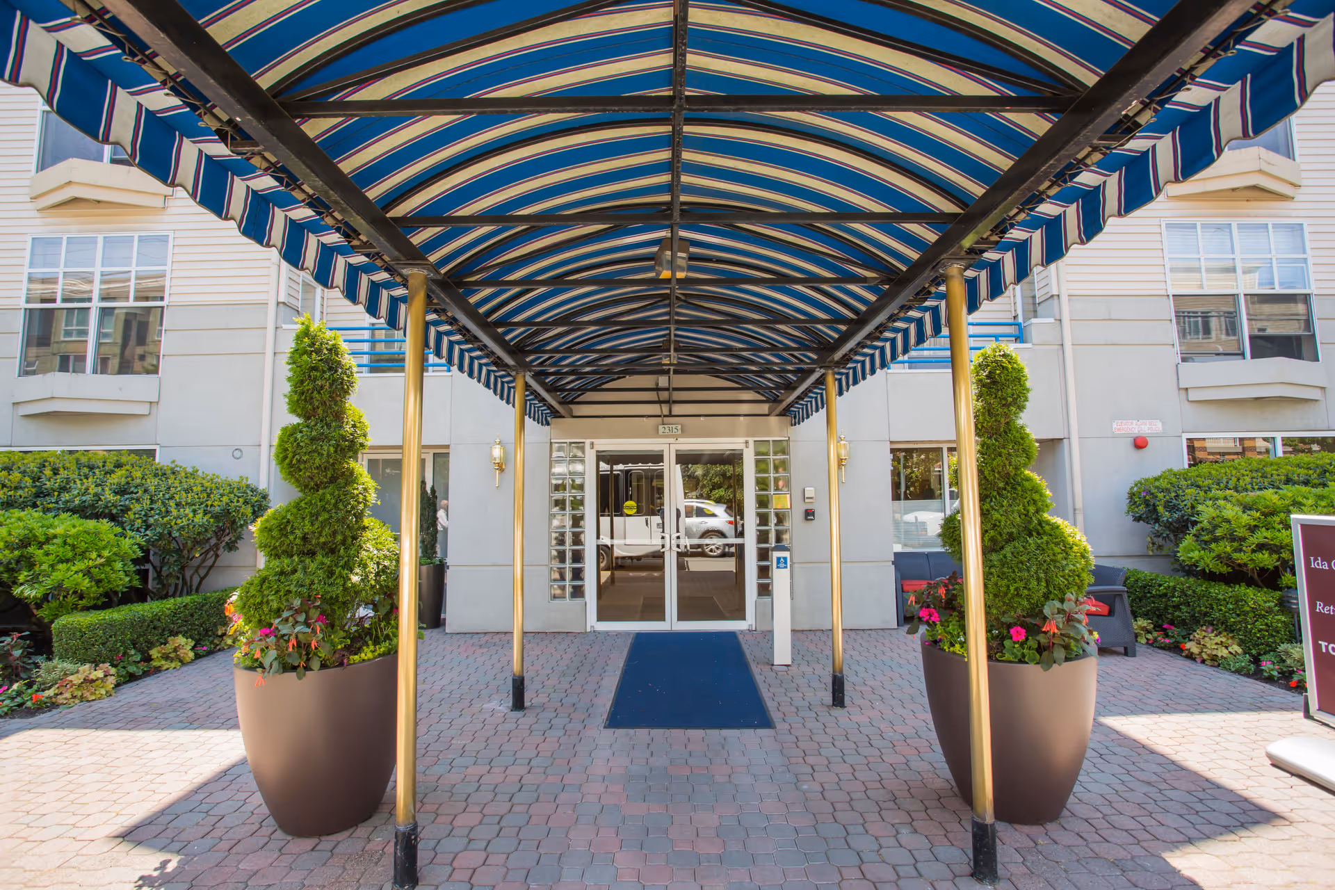 Entrance to a building with a blue and white striped canopy supported by gold poles. Two large planters with spiral-shaped topiary plants and flowers flank the walkway leading to glass double doors. The building exterior is light-colored with windows and greenery on either side.