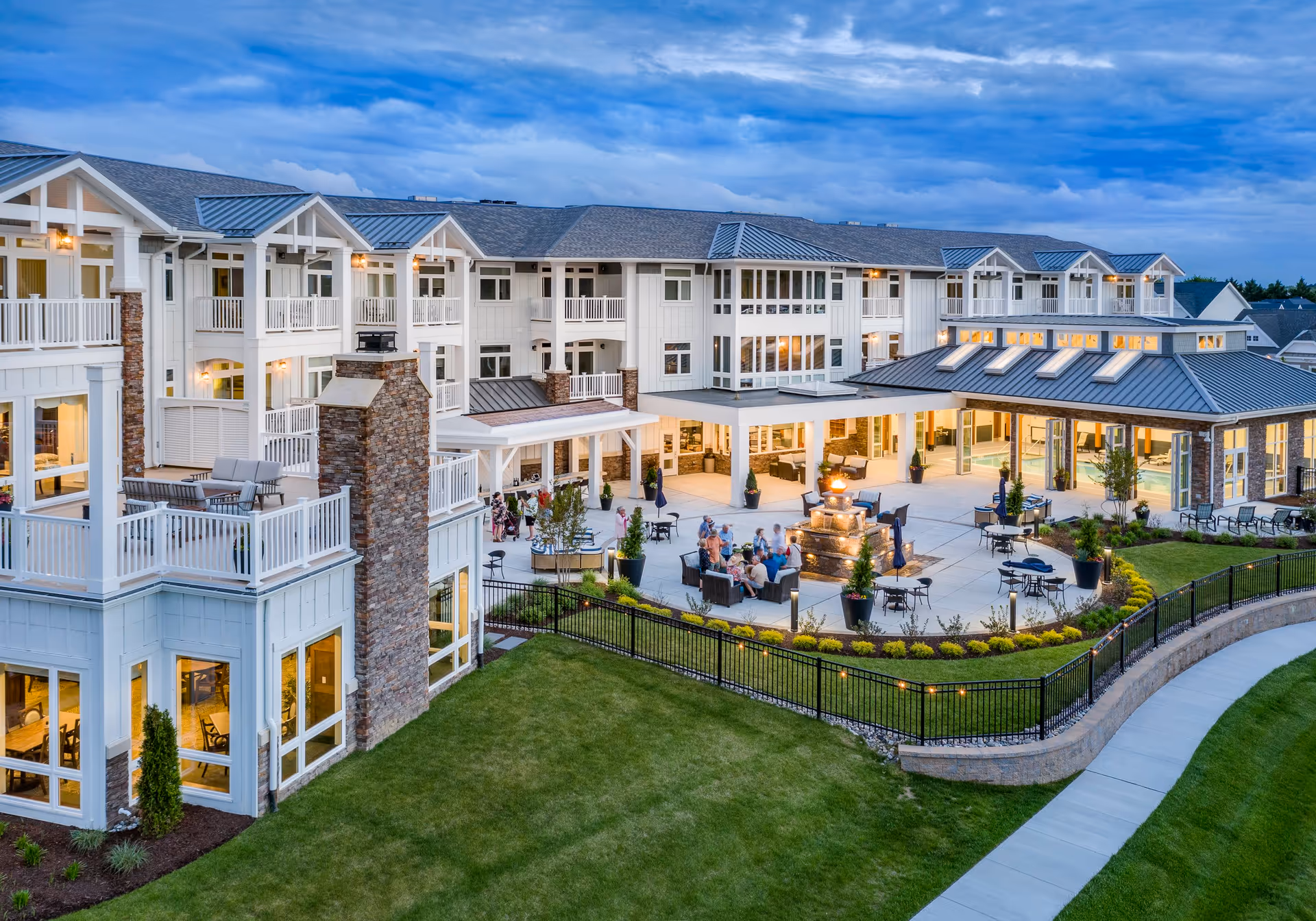 Evening aerial view of The Lodge at Truitt Homestead senior living facility showing a large, multi-story building with balconies and outdoor seating areas. Residents are gathered around a central fire pit on a spacious patio surrounded by well-maintained landscaping and a curved walkway.