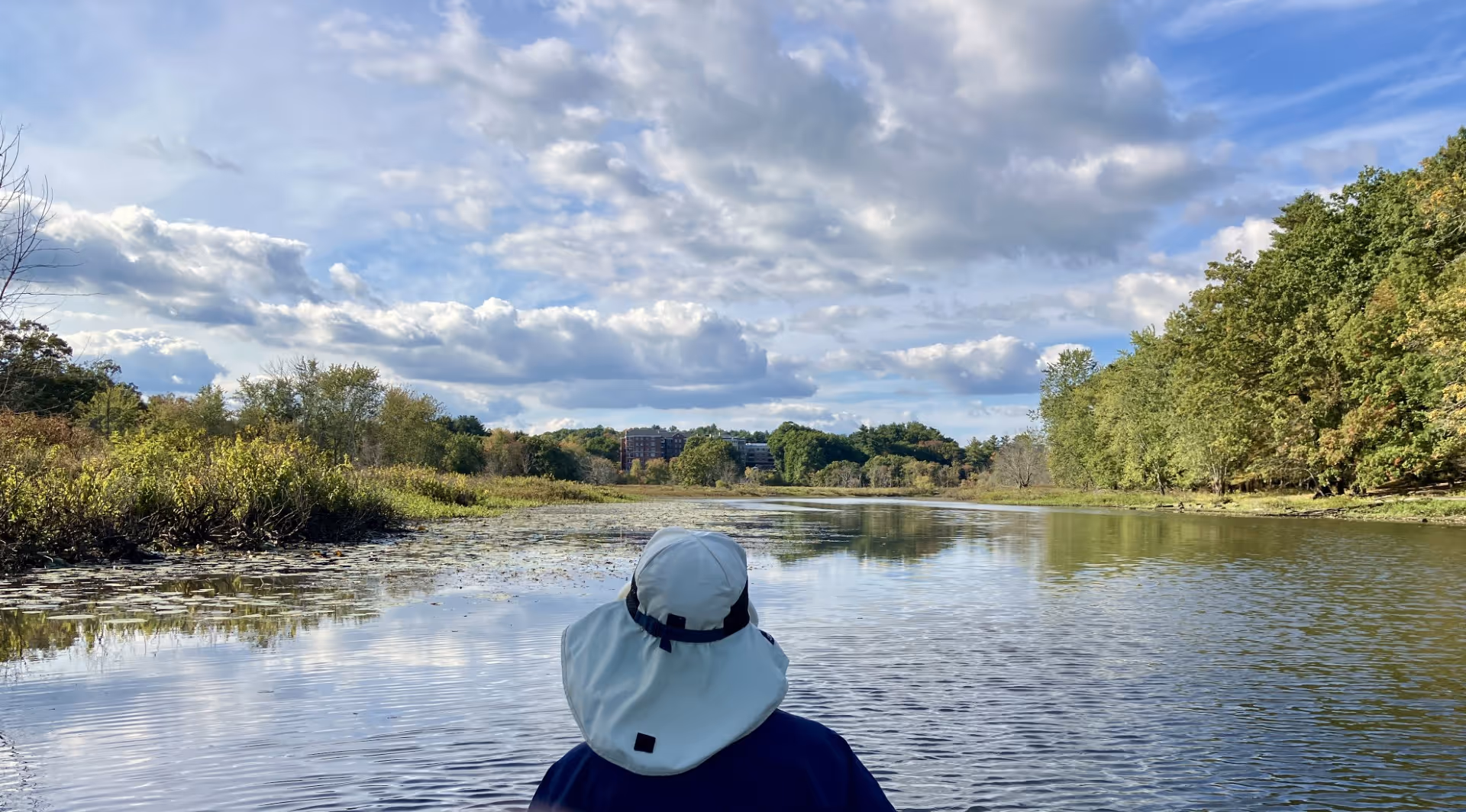 View from behind a person wearing a wide-brimmed hat looking out over a calm river surrounded by green trees and vegetation under a partly cloudy sky.