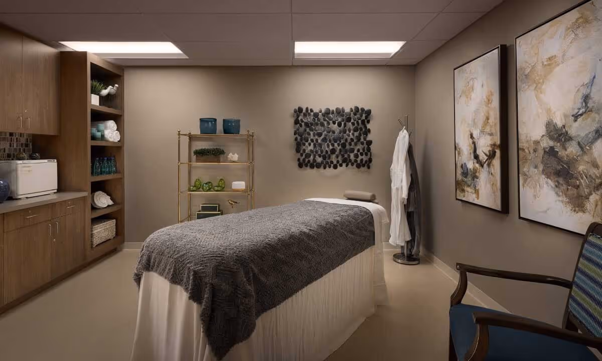 A calming treatment room with a massage table draped in a gray blanket, shelving and cabinets, wall art, and a robe on a stand.