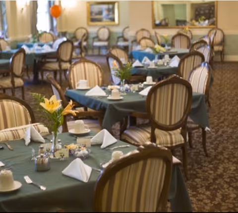 A dining room with multiple tables covered in green tablecloths, each set with white napkins, cups, and silverware. The chairs have wooden frames with striped upholstery. There are flower arrangements on the tables and framed artwork on the walls.