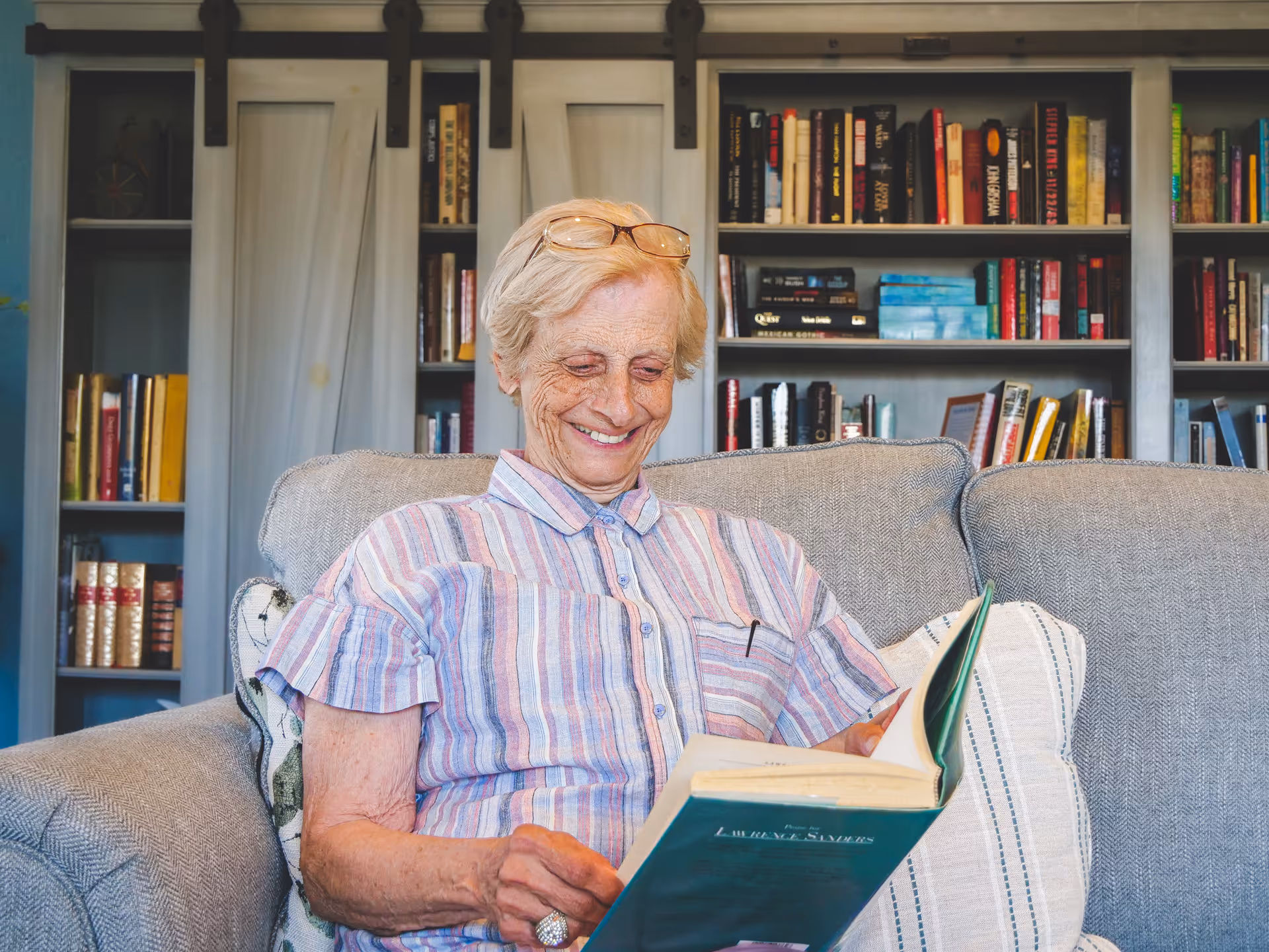 An elderly woman with short gray hair and glasses resting on her head is sitting on a gray couch, smiling while reading a book. Behind her is a bookshelf filled with various books.