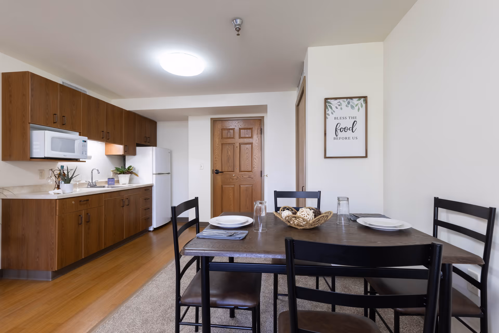 A kitchen and dining area in a senior living facility with wooden cabinets, a white microwave, refrigerator, and a sink. A wooden dining table with four black chairs is set with plates, glasses, and napkins. A framed sign on the wall reads 'Bless the food before us.'