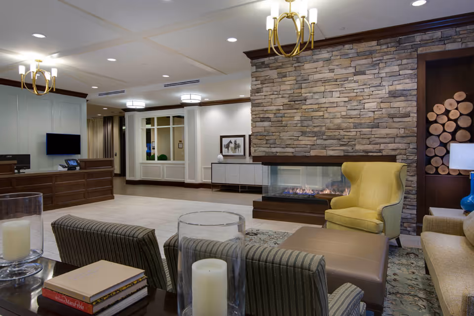 A cozy assisted living facility common area featuring a stone fireplace with a fire burning, a yellow armchair, a beige sofa, striped chairs, and a coffee table with candles and books. The room has warm lighting with chandeliers and recessed ceiling lights, a wooden reception desk with a computer and phone, and decorative wood logs stacked in a niche beside the fireplace.