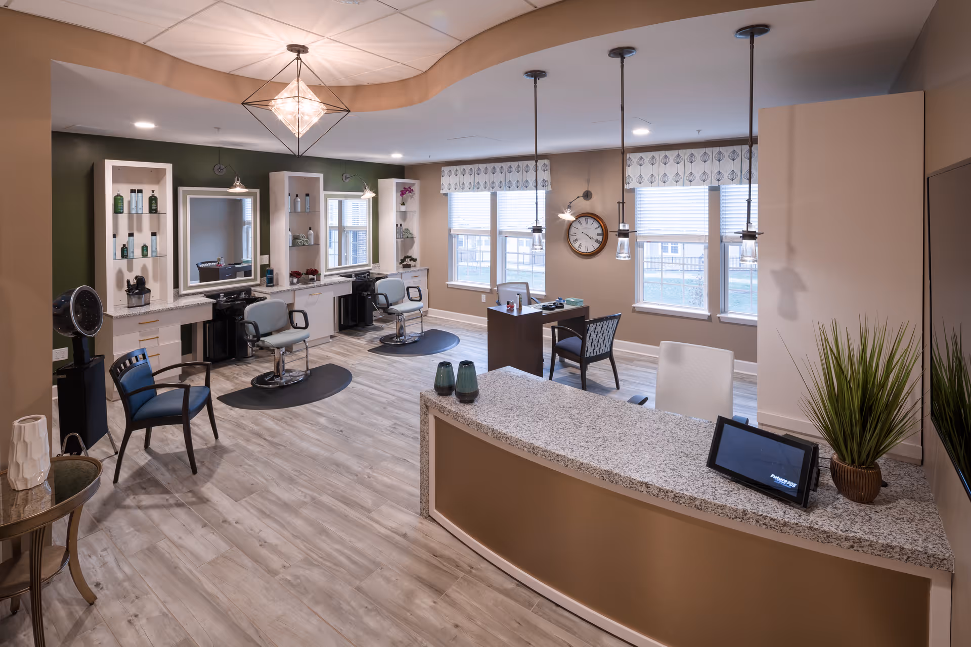 Interior view of a modern salon area in a senior living facility with two styling chairs in front of mirrors and shelves with hair products, a reception desk with a tablet and decorative plants, a small desk with chairs near windows, and wooden flooring.