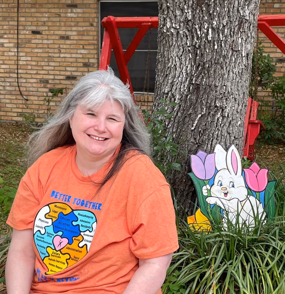 A smiling woman with long gray hair wearing an orange t-shirt with a colorful puzzle heart design sits outdoors in front of a large tree. Behind her, there is a red wooden swing and a decorative cutout of a white bunny with tulips in the grass. A brick building wall is visible in the background.