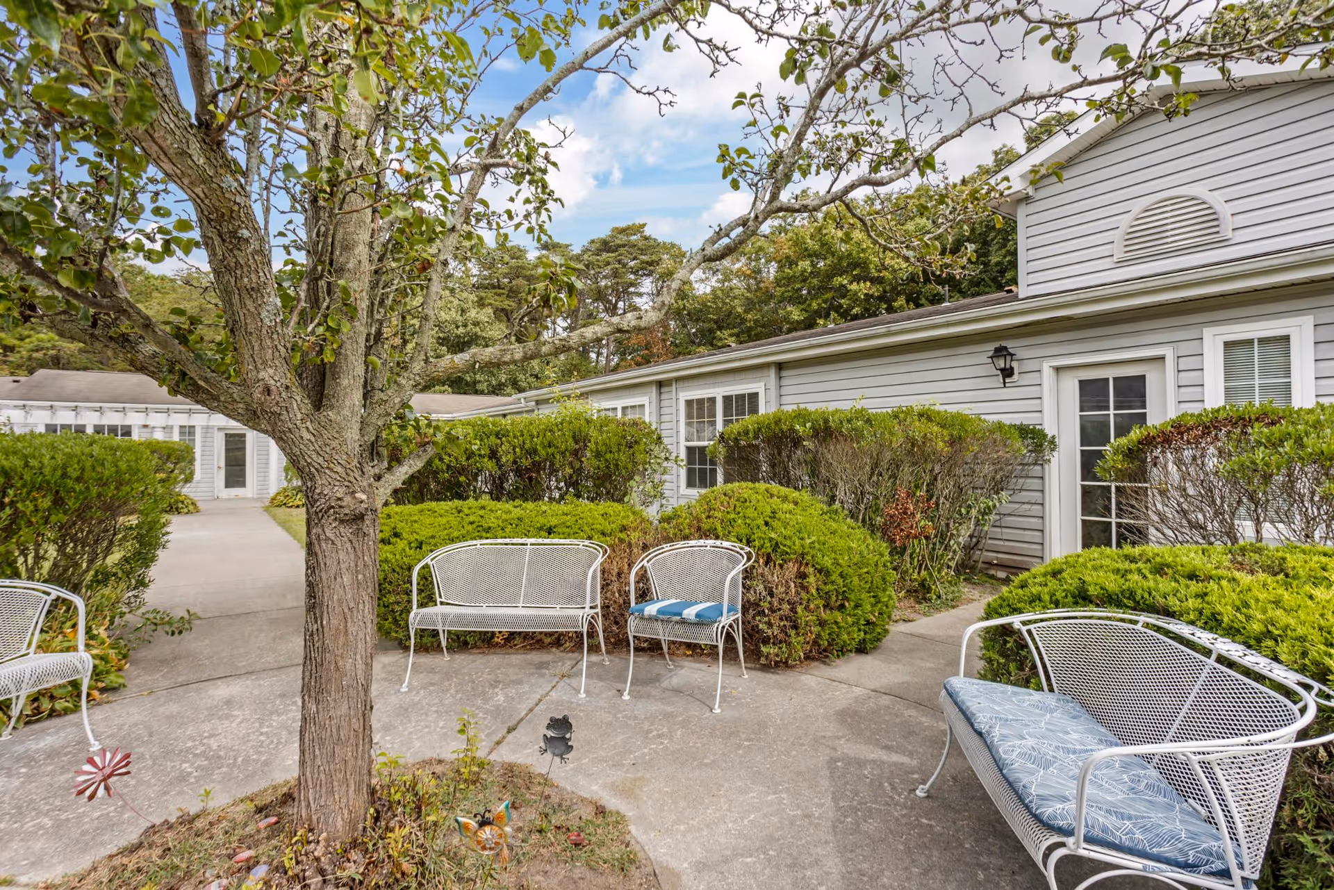 Outdoor seating area at The Addison of Somers Place featuring white metal benches with cushions arranged around a tree, surrounded by green bushes and a gray building with white-framed windows and doors in the background under a partly cloudy sky.