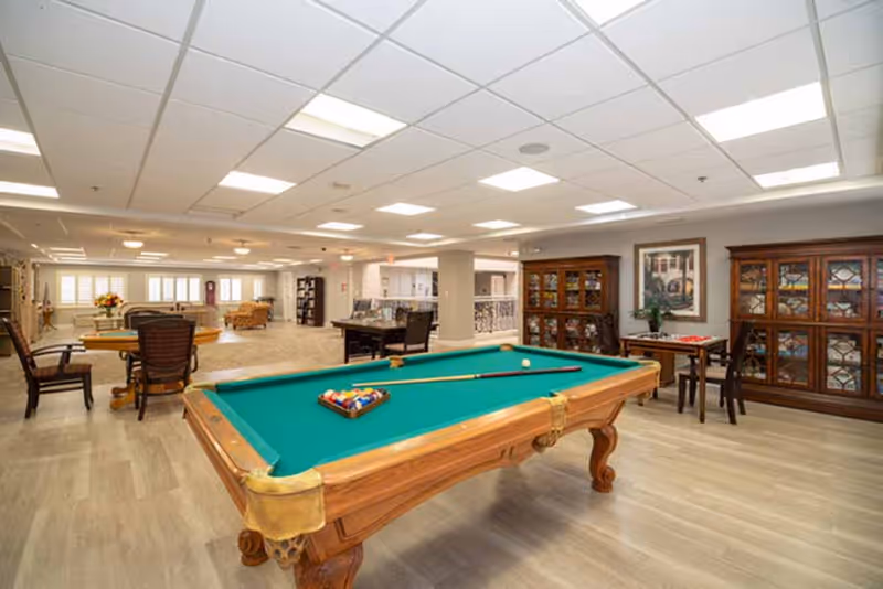 A spacious recreational room with a pool table in the foreground, several chairs and tables scattered throughout, wooden display cabinets against the wall, and bright overhead lighting in a senior living facility.