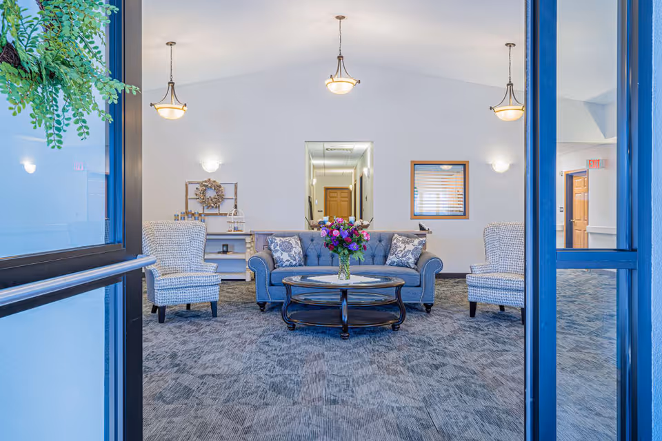 View through glass doors into a bright, spacious living room area with a gray sofa adorned with patterned pillows, two matching armchairs, and a round wooden coffee table with a vase of colorful flowers. The room has light walls, carpeted floor, three hanging ceiling lights, and decorative wall sconces. A hallway and a window with blinds are visible in the background.