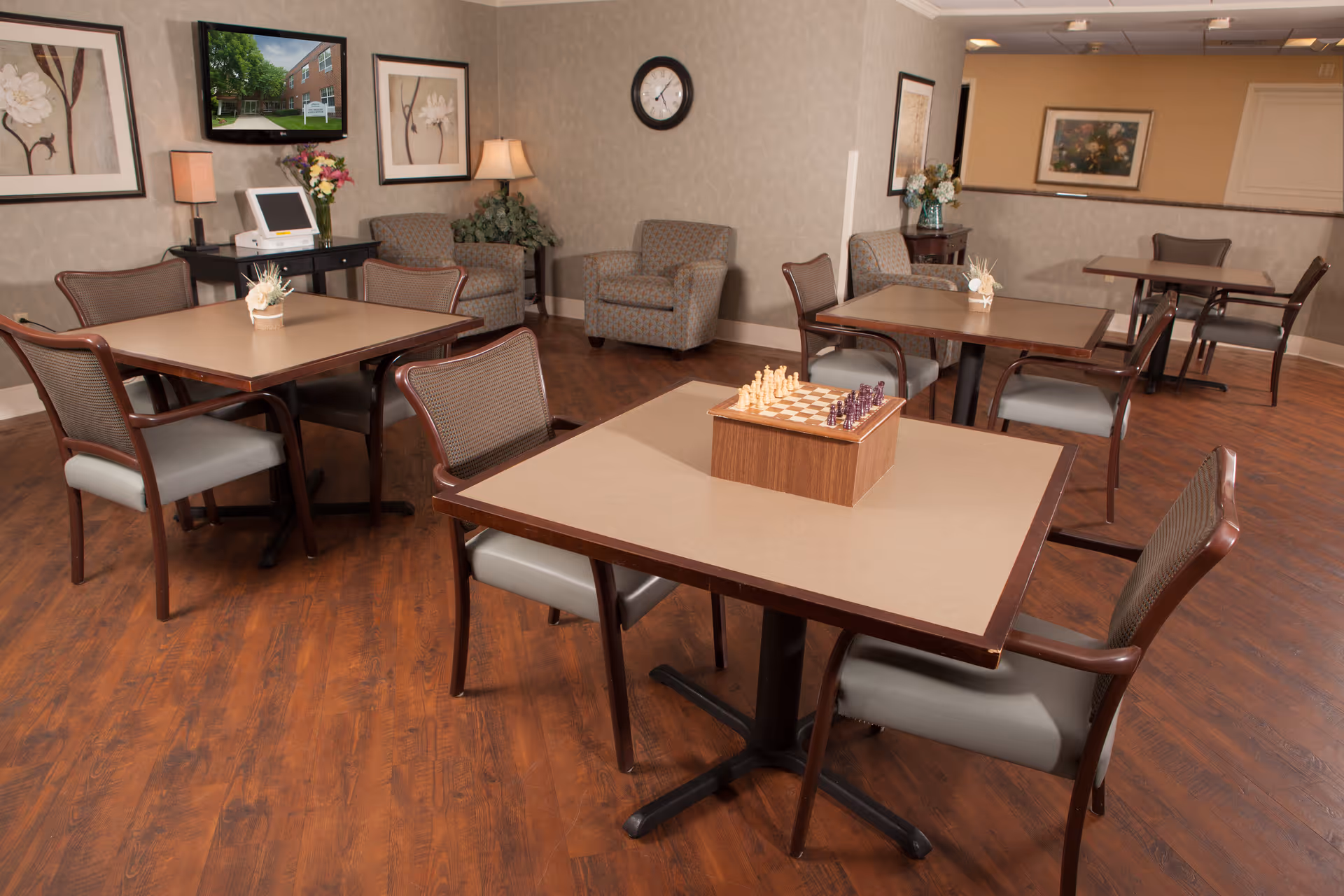 Community dining/activity room with multiple tables and chairs, a chess set on a table, a wall-mounted TV and lounge chairs in the background.