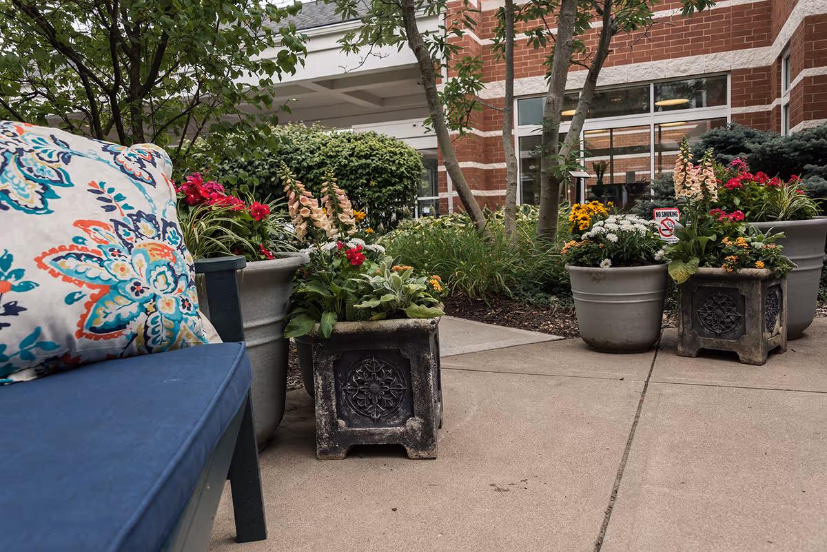 Outdoor patio area with a blue cushioned bench featuring a colorful floral pillow, surrounded by multiple flower pots containing various blooming flowers and greenery. The background shows part of a brick building with large windows and some trees.
