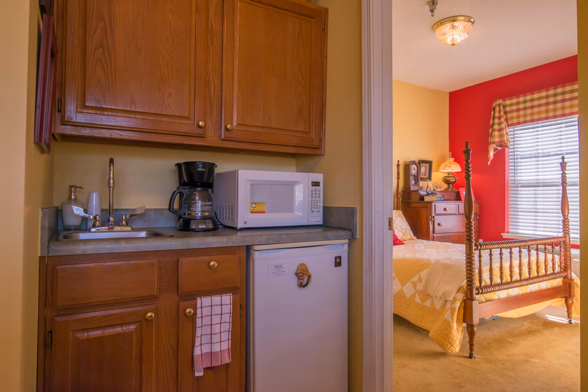 A small kitchenette area with wooden cabinets, a sink, a coffee maker, a microwave, and a mini refrigerator. To the right, an open doorway reveals a bedroom with a wooden four-poster bed, a dresser, a lamp, and a window with blinds and a plaid valance.