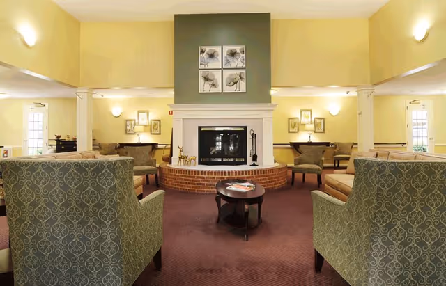 A cozy living room area in a senior living facility featuring a central fireplace with a brick base and white mantel. Above the fireplace are four framed floral artworks. The room is furnished with patterned armchairs and sofas arranged around a dark wooden coffee table with magazines. The walls are painted light yellow, and there are wall-mounted lights providing warm illumination. Doors and windows are visible on either side of the room.