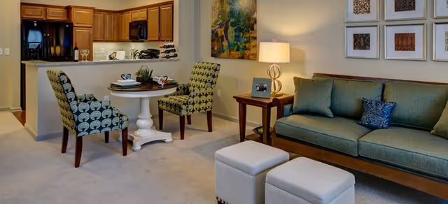 A cozy living area featuring a green sofa with cushions, two white ottomans, a wooden side table with a lamp and framed photo, and a small round dining table with two patterned chairs. The background shows a kitchen with wooden cabinets and black appliances, and colorful artwork on the walls.