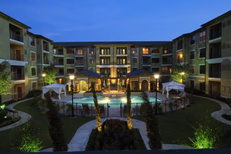 Night view of a multi-story residential building surrounding a central courtyard with a swimming pool, illuminated street lamps, and seating areas with umbrellas.