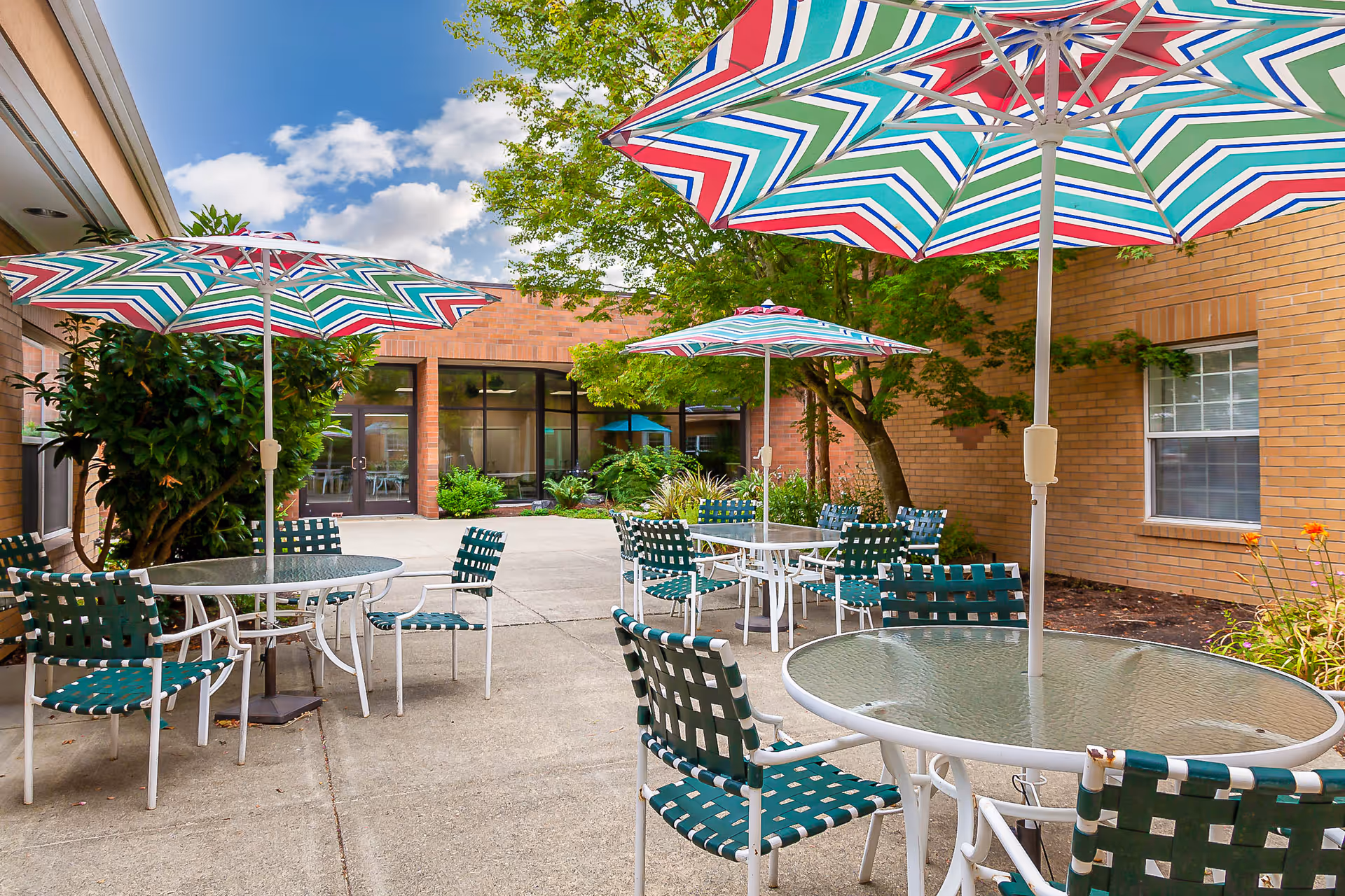 Outdoor patio area with several round glass tables and green woven chairs. Each table has a large colorful striped umbrella providing shade. The patio is surrounded by brick walls, windows, and some greenery including trees and plants. The sky is partly cloudy and blue.