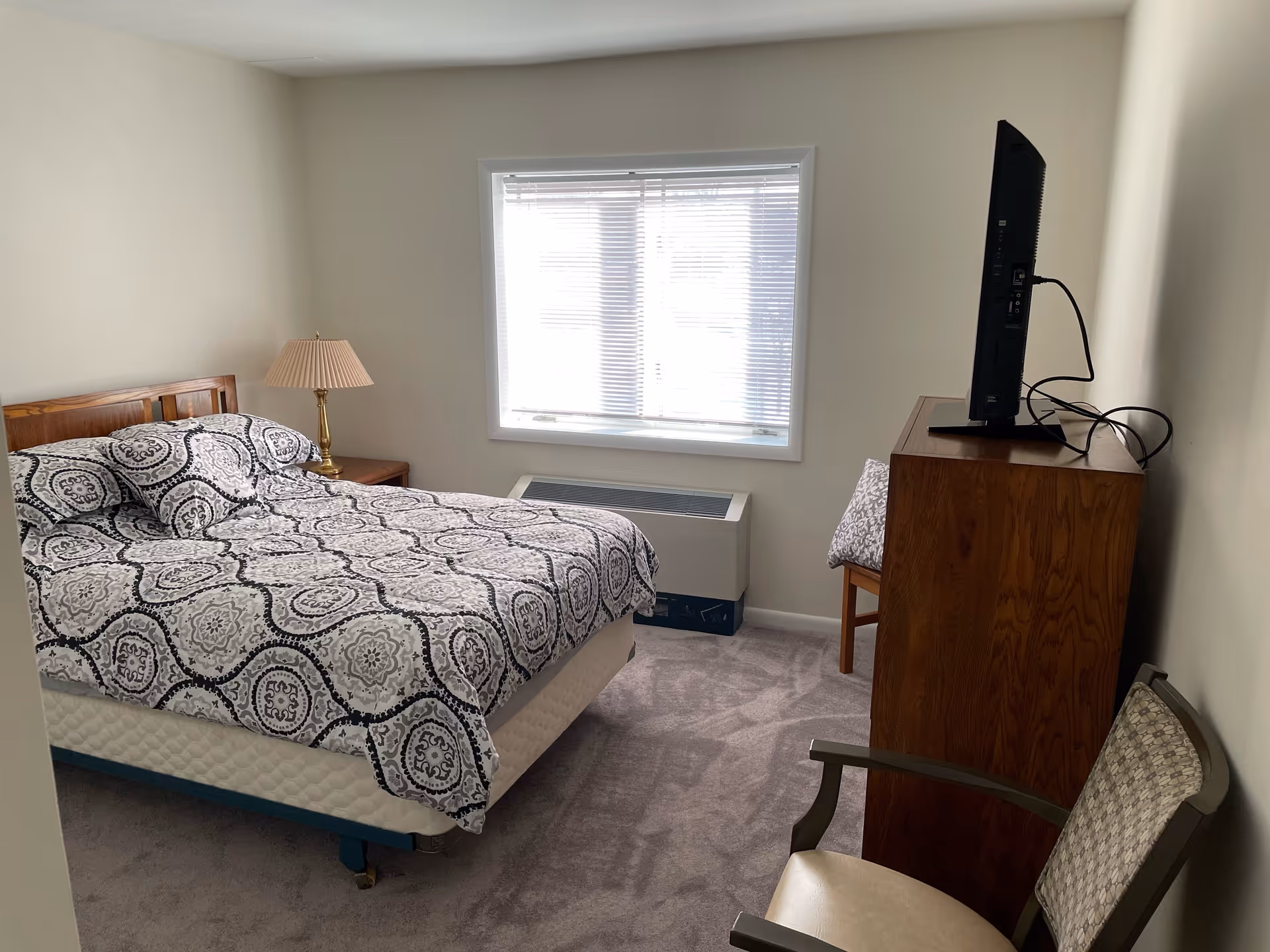 A simple bedroom with a bed covered in a patterned black and white bedspread, a wooden headboard, a bedside table with a lamp, a window with blinds, a wooden dresser with a flat-screen TV on top, and a chair with a cushioned seat and backrest.