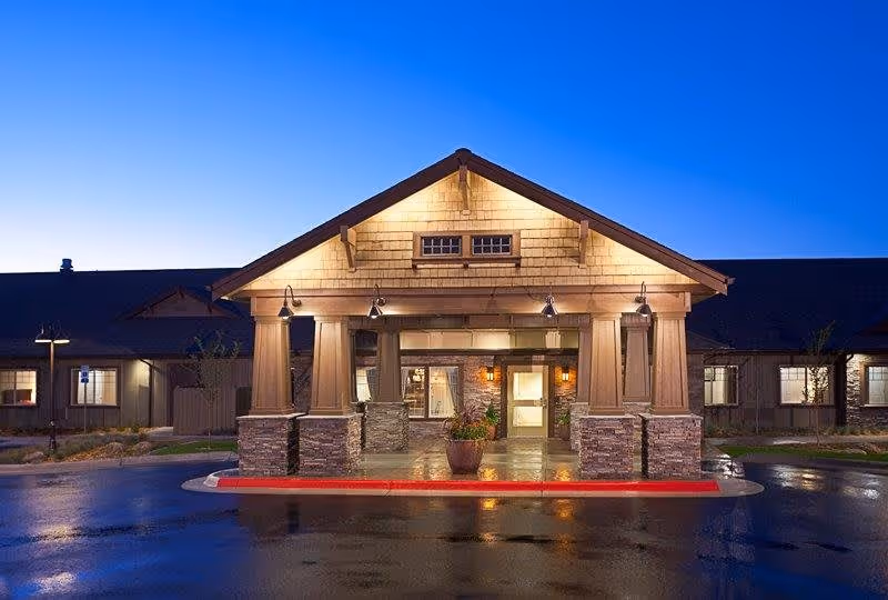 Exterior front entrance of Canyon Creek Memory Care Community at dusk, featuring a covered driveway with stone pillars and warm lighting under a peaked roof.
