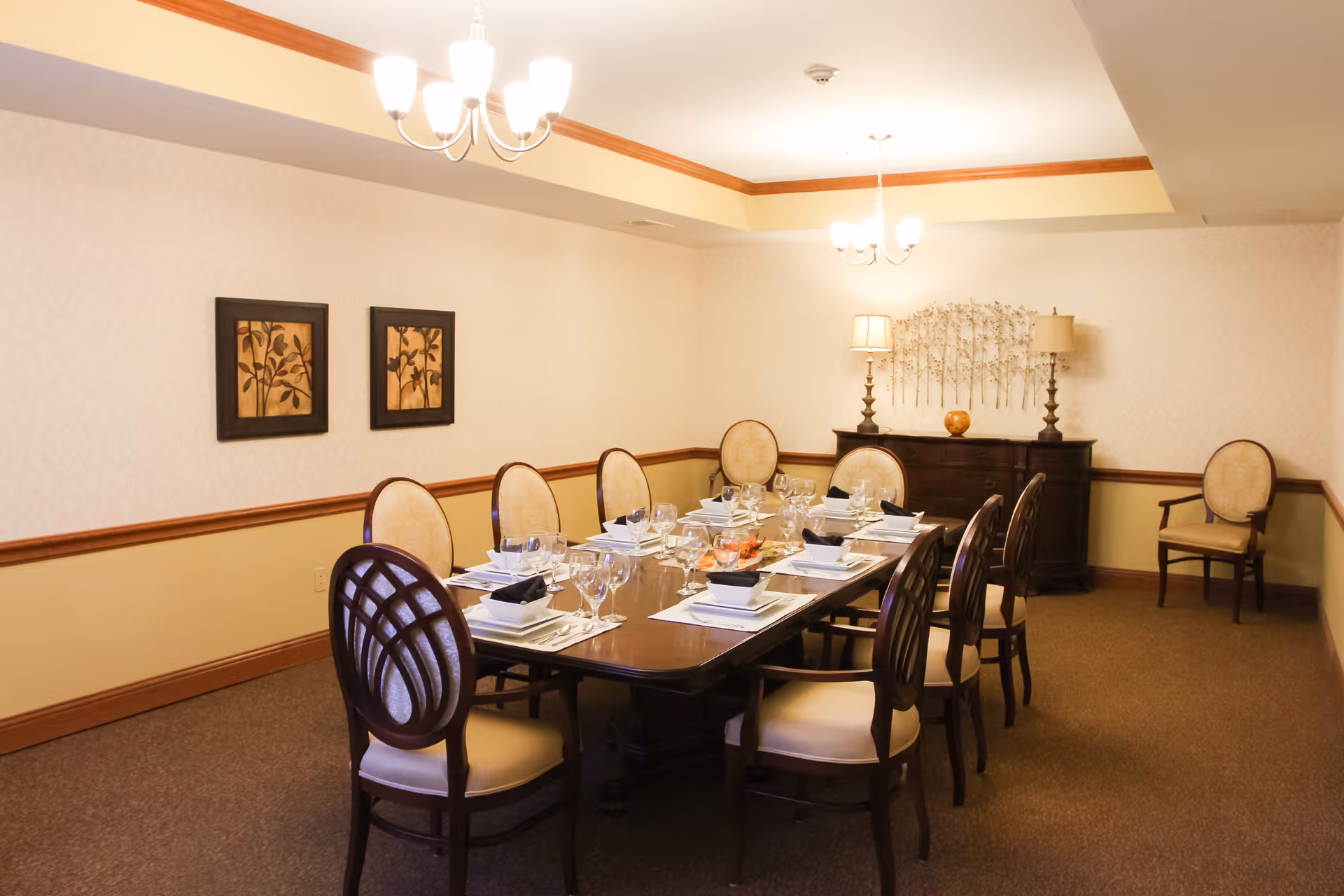 A formal dining room with a long wooden table set for eight people. The table is arranged with white plates, bowls, black napkins, and glassware. The room has beige walls with a wooden chair rail and two framed botanical prints on one wall. There are two chandeliers hanging from the ceiling and a dark wooden sideboard with two lamps and decorative items against the far wall. Additional chairs are placed along the walls.