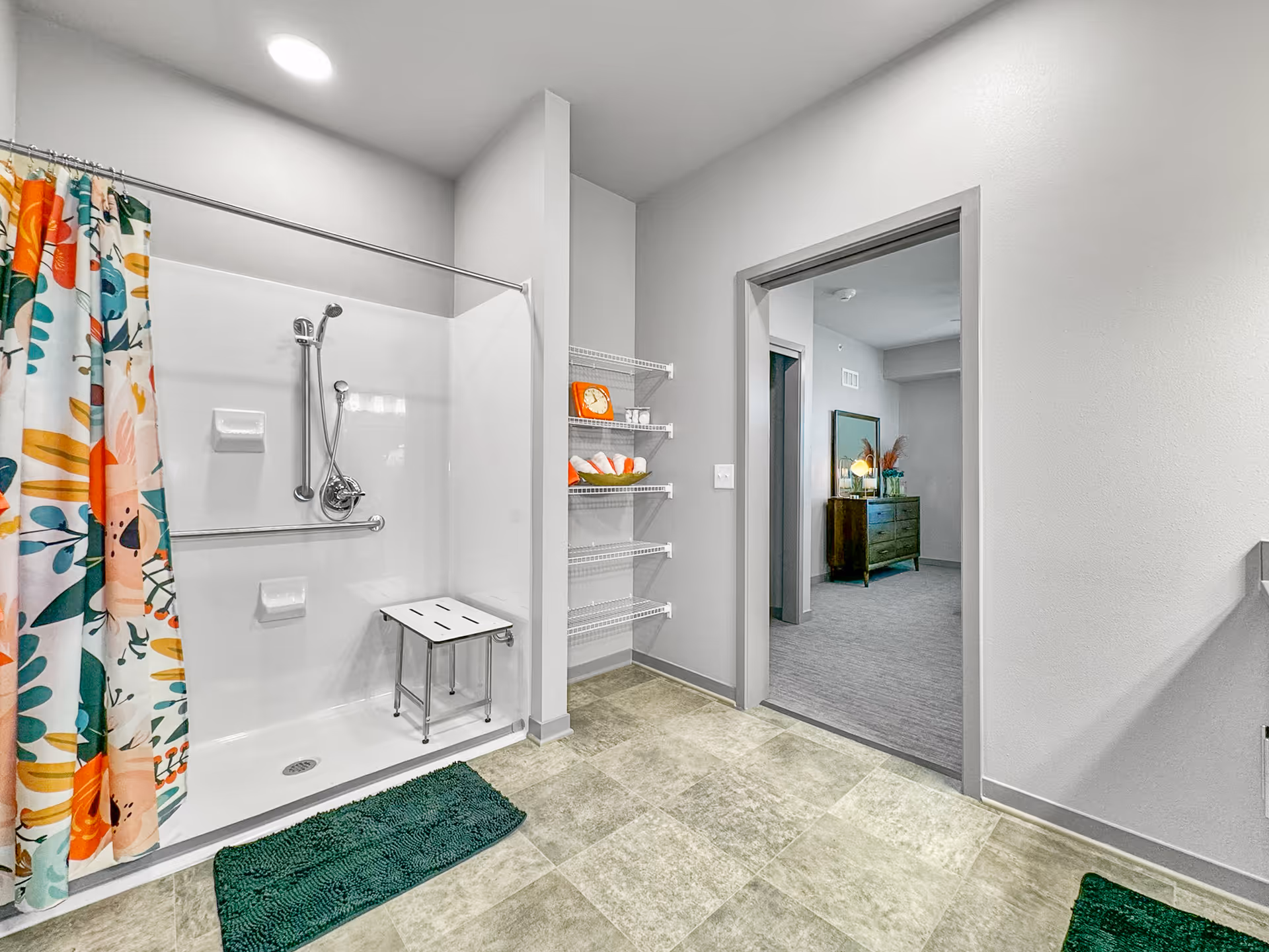 A bathroom with a walk-in shower featuring a colorful floral shower curtain, a white shower seat, and grab bars. There are wire shelves holding rolled towels and an orange clock. The bathroom floor is tiled, and an open doorway leads to a carpeted room with a dresser and a lamp.