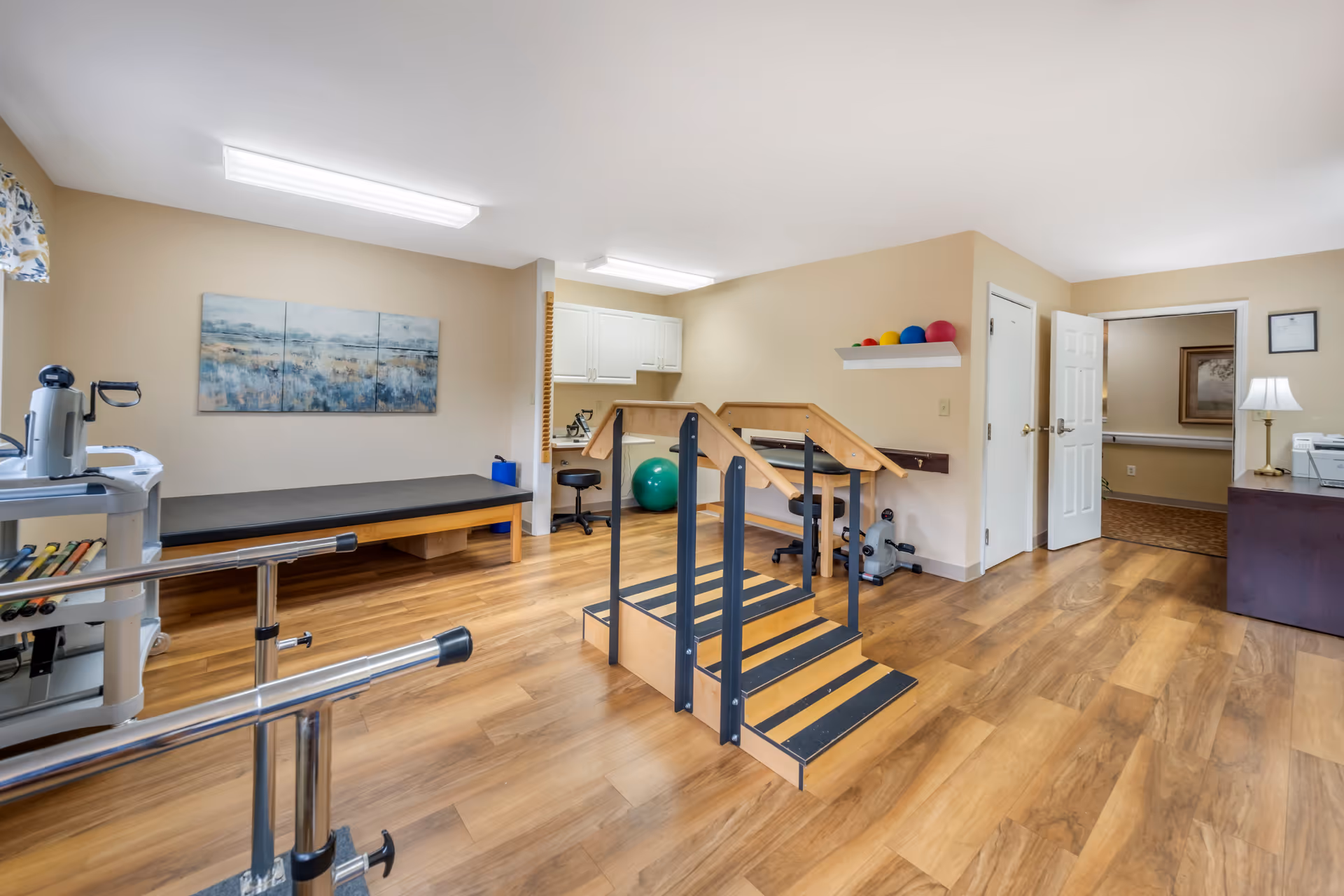 A physical therapy or rehabilitation room with wooden flooring, a small set of stairs with handrails for practice, exercise balls on a shelf, a padded therapy table, and various therapy equipment. The room has beige walls, fluorescent ceiling lights, and a doorway leading to another room.