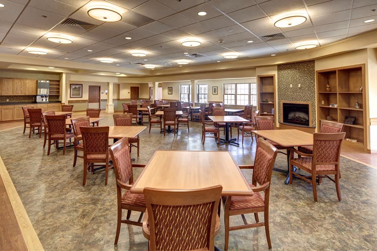 A spacious assisted living dining room with multiple wooden tables and chairs arranged neatly. The room features large windows allowing natural light, a fireplace with a decorative tile surround, built-in wooden shelves, and a kitchenette area with cabinets and a microwave. The ceiling has recessed lighting and circular light fixtures.